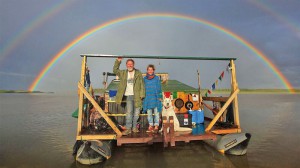 Foto von Mario und Ramona Goldstein mit ihrem Hund auf dem selbstgebauten Floß. Im Hintergrund ein doppelter Regenbogen