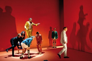 Szenenbild mit sieben Schauspielern aus "Caligula" im Theater Basel. Manche tragen keine Hosen, ihre Körper werfen große Schatten auf die rote Wand im Hintergrund