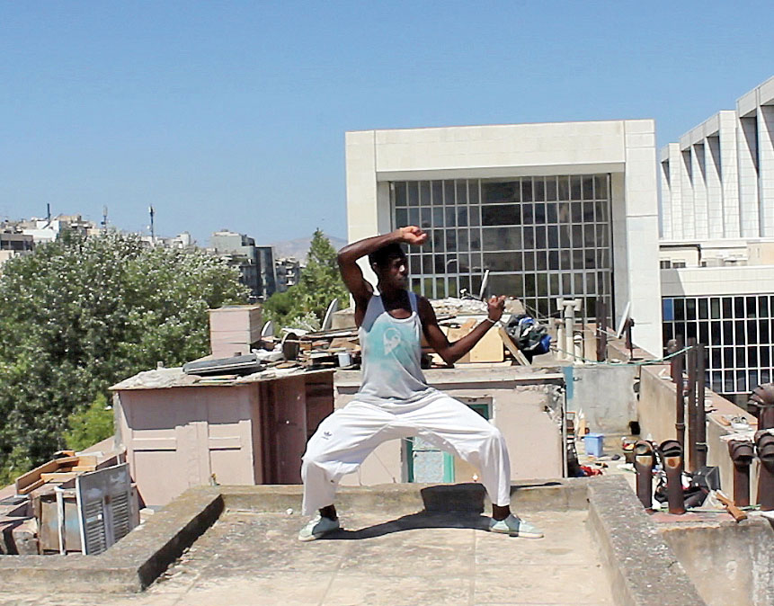 Filmstill "Research Refugees". Jenseits der gängigen Fernsehbilder. © Therese Koppe Foto eines dunkelhäutigen Mannes auf einem Hausdach. Er vollführt eine Bewegung die an Tai-Chi oder Yoga erinnert.